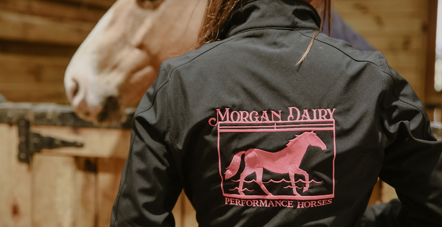 Una imagen de primer plano de una chaqueta negra con costuras rosas, con las palabras “Morgan Dairy Performance Horses”, usada por una mujer con cabello largo, liso y castaño.